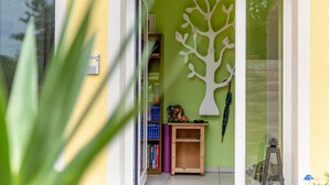 Una stanza con una parete verde, una decorazione a forma di albero bianco, uno scaffale di legno con libri e una porta vetrata che dà sull'esterno.