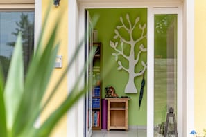 A room with a green wall, a white tree decoration, a wooden shelf with books, and a glass door leading outside.