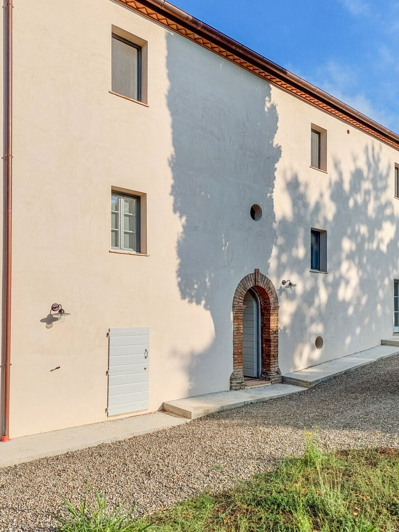 A building with a light-colored facade, a small arched entrance, and a gravel driveway.