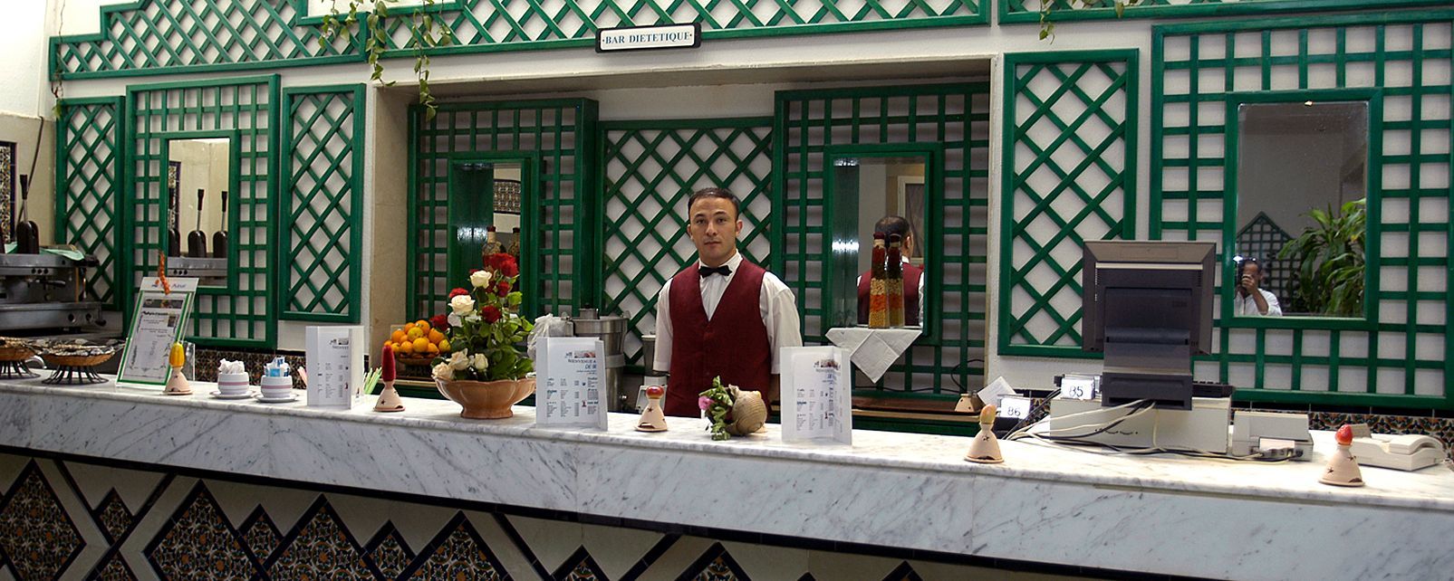 Un bar orné d’un treillis vert, un comptoir où sont exposés divers produits, et une enseigne indiquant « BAR DIÉTÉTIQUE ».