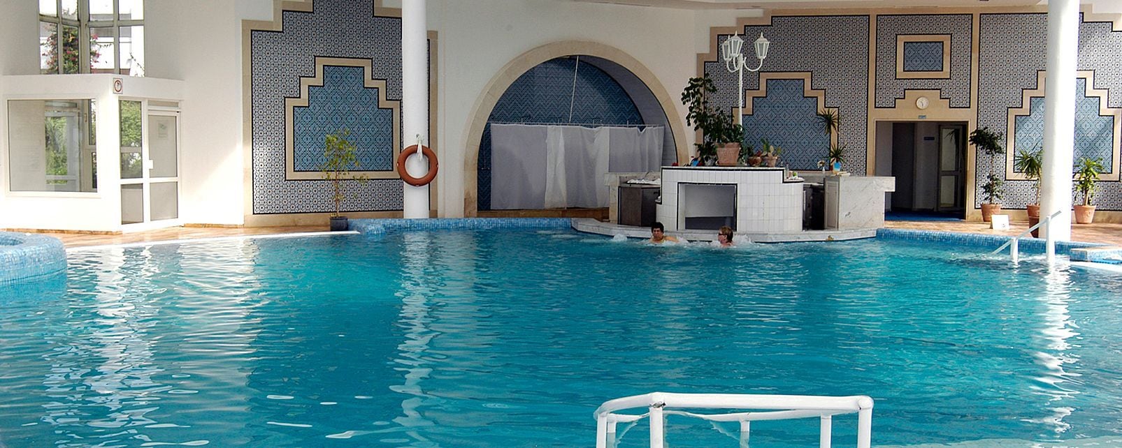 An indoor swimming pool with clear blue water, a lifeguard stand, and a reception area with a counter and plants.