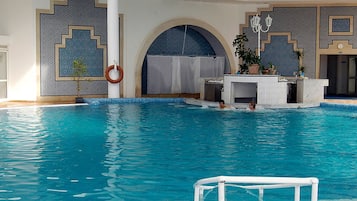 An indoor swimming pool with clear blue water, a lifeguard stand, and a reception area with a counter and plants.