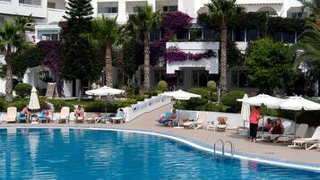 A pool area with lounge chairs and umbrellas, surrounded by palm trees and a building with balconies.