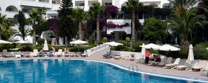 A pool area with lounge chairs and umbrellas, surrounded by palm trees and a building with balconies. - Royal Azur Thalassa (Hammamet)