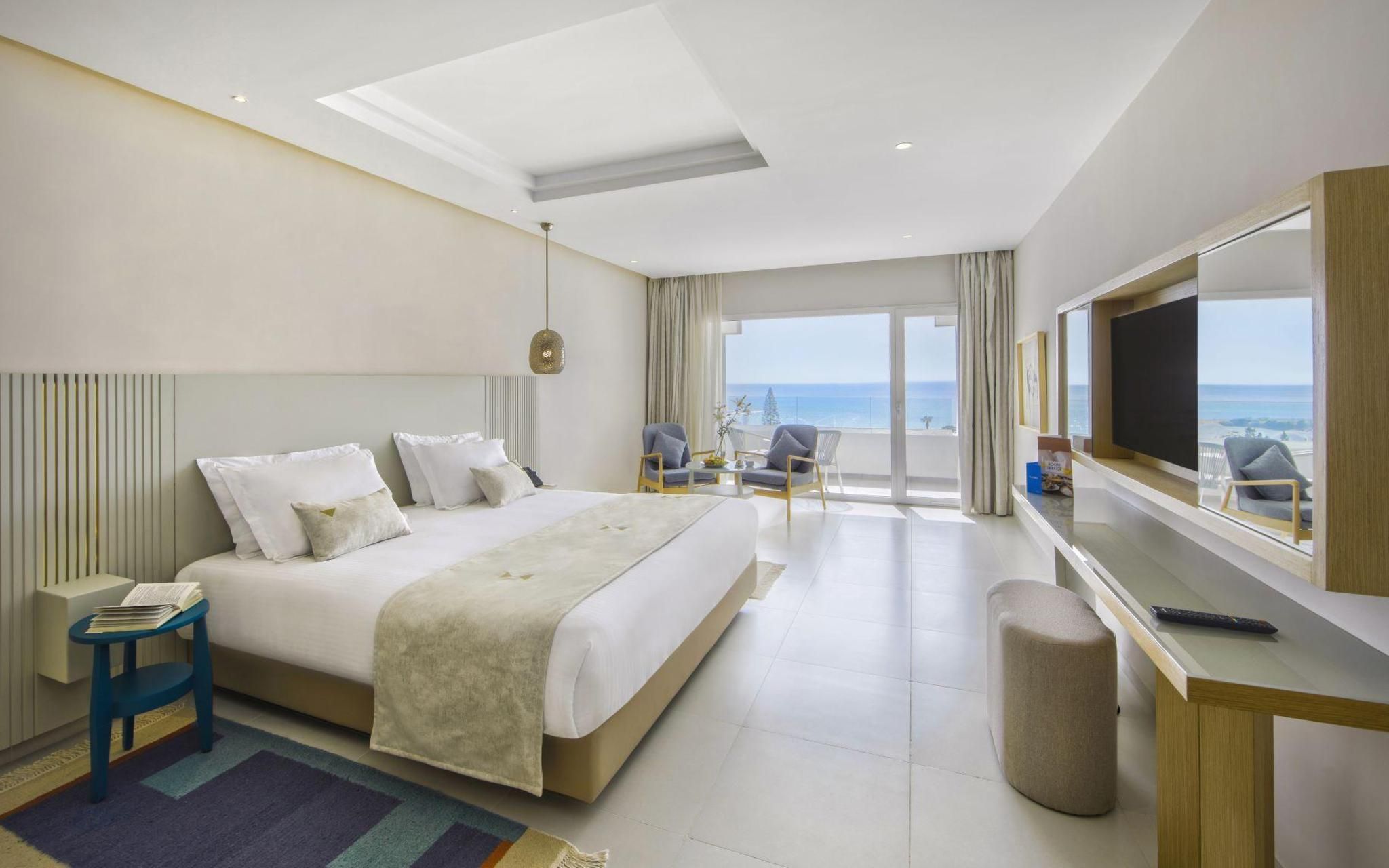 Une chambre d’hôtel comprenant un lit, un bureau, une chaise, une télévision et un balcon avec vue sur la mer.