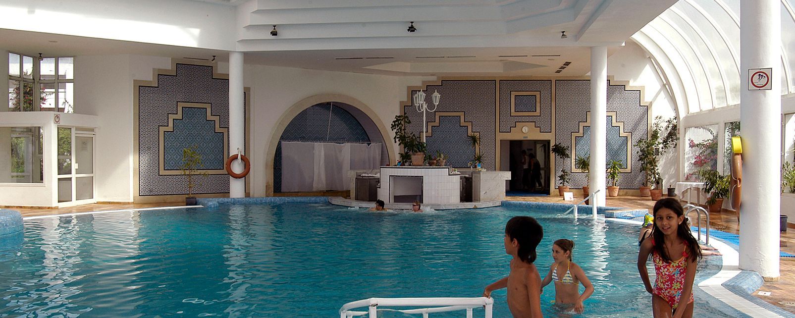 Une piscine intérieure dotée d’un bar central, entourée de murs recouverts de carreaux aux motifs géométriques.