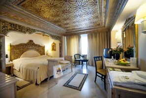 A hotel room with a large bed, a desk, a chair, a television, and a dining table with a vase of flowers. - Royal Azur Thalasso Golf (Hammamet)