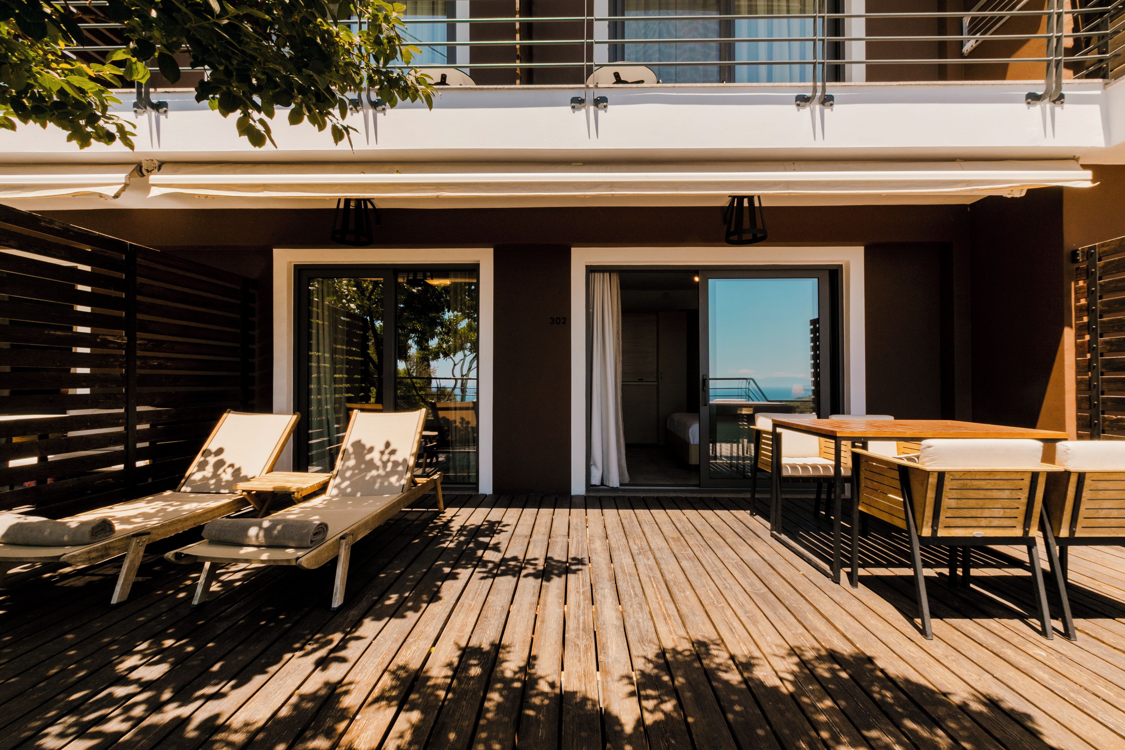 A wooden deck with lounge chairs and a table, a balcony with a railing, and a view of the sea.