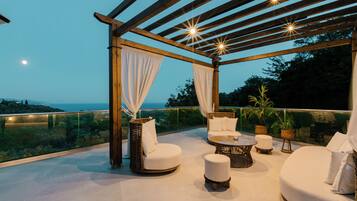 A covered outdoor seating area with wicker furniture, a round coffee table, and a view of the sea.
