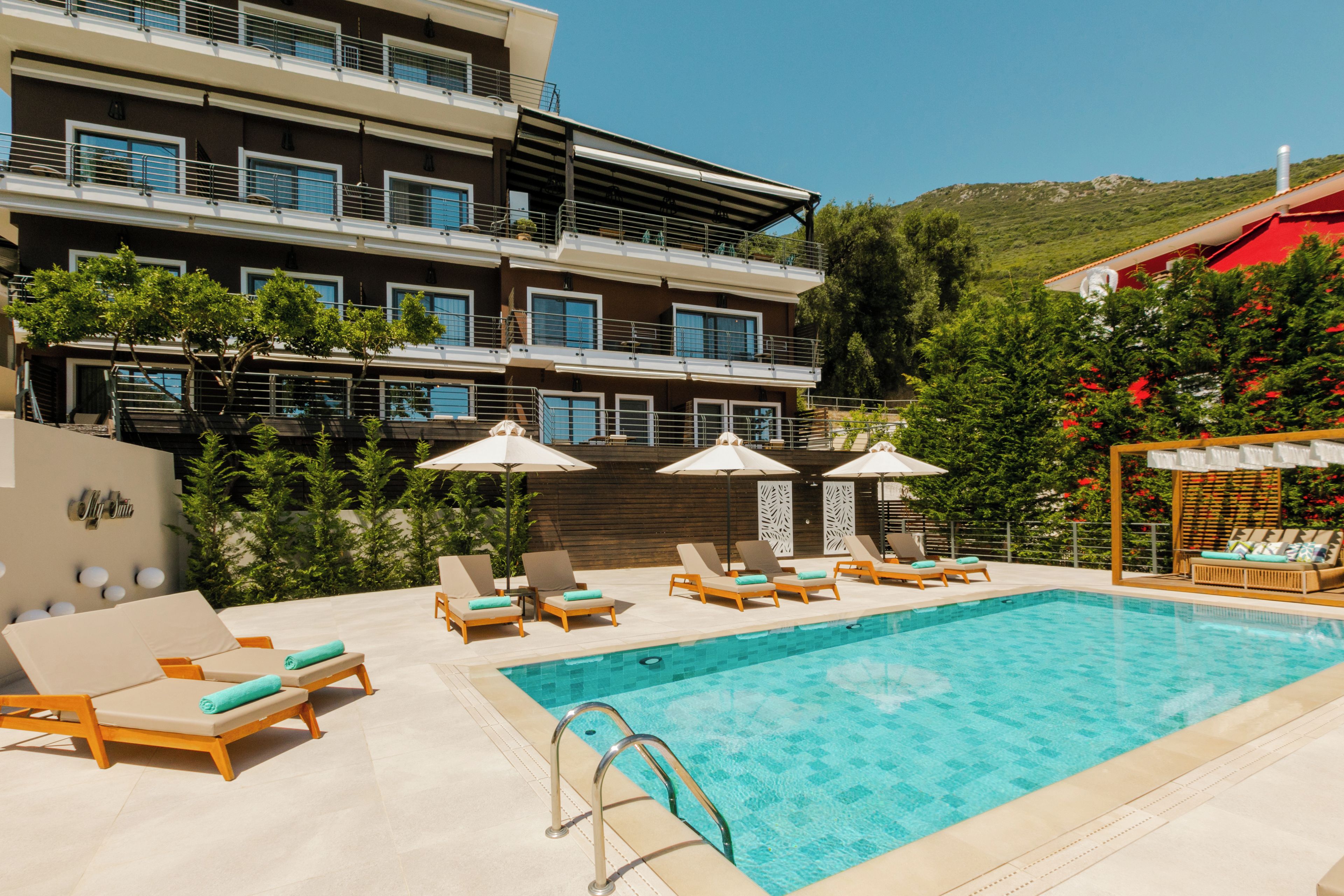 Una piscina con sillones de descanso y sombrillas, un edificio de varios pisos con balcones, y una colina al fondo.