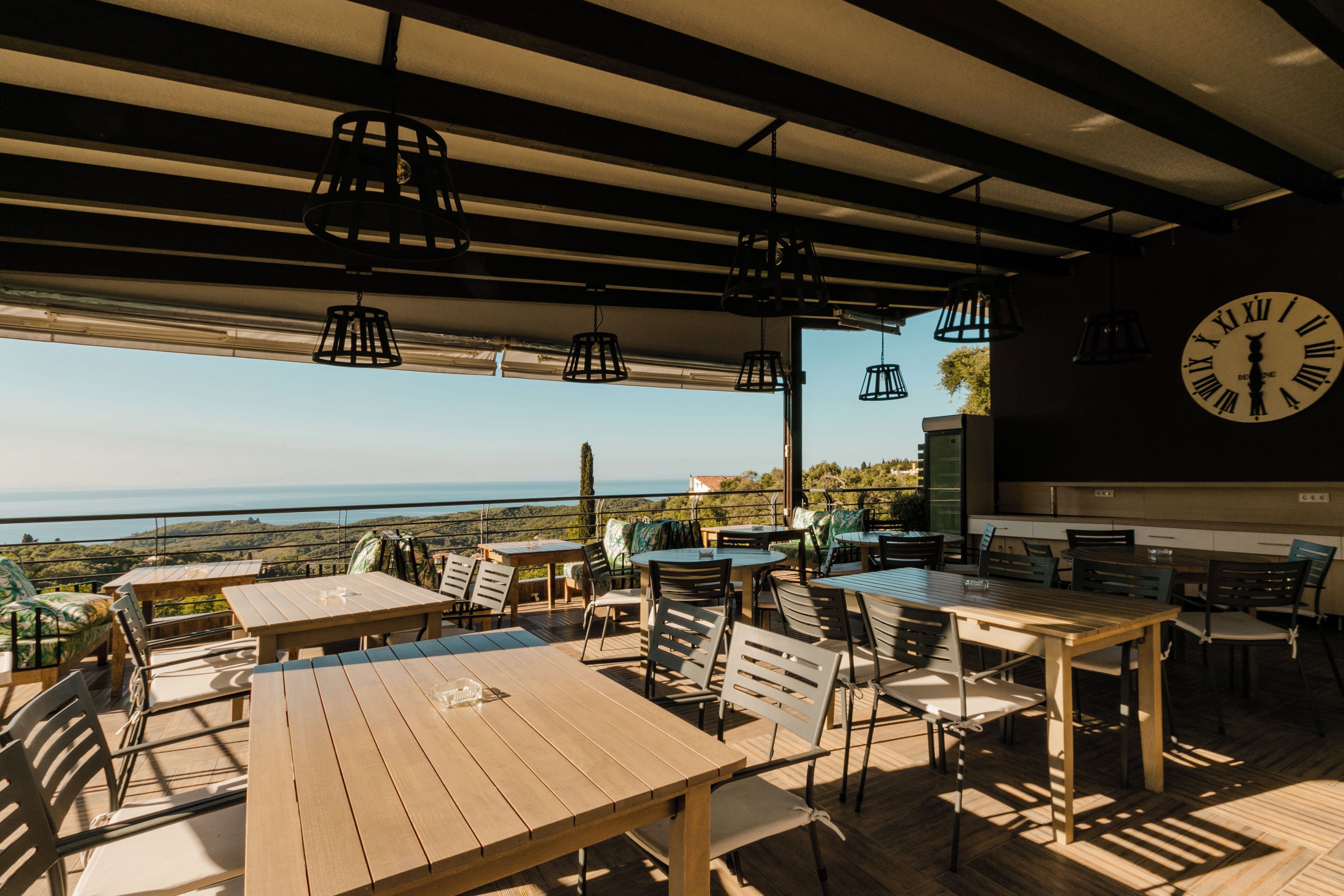 An outdoor dining area with tables and chairs, a large clock on the wall, and a view of the landscape.
