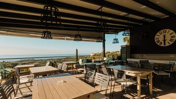 An outdoor dining area with tables and chairs, a large clock on the wall, and a view of the landscape.