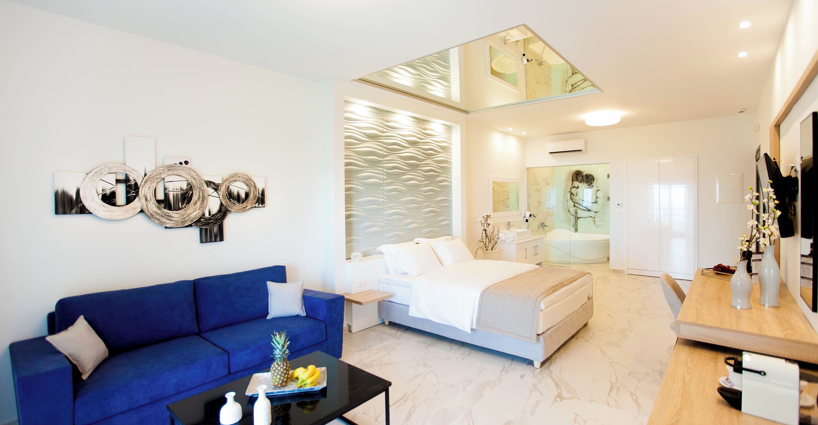 Una habitación de hotel moderna con un sofá azul, una cama, una mesita con fruta, un espejo y un baño con bañera y ducha.