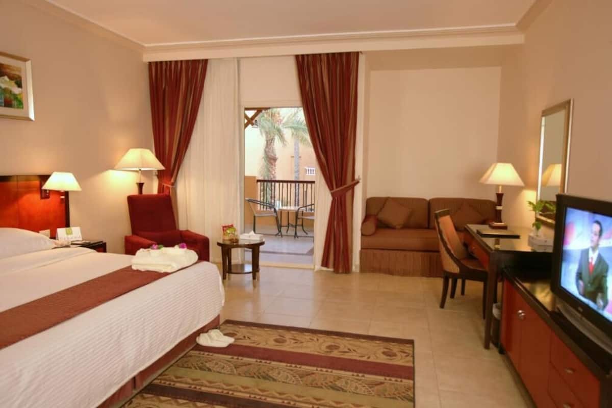 Habitación de hotel con cama, televisión, escritorio, silla, sofá y balcón con cortinas.