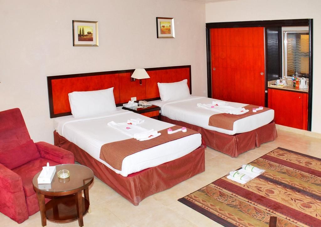 A hotel room with two beds, a red chair, a small table, and a red wardrobe.