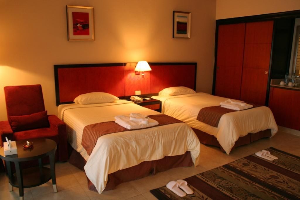 A hotel room with two beds, a red armchair, a small table, and a nightstand with a lamp.