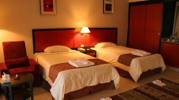 A hotel room with two beds, a red armchair, a small table, and a nightstand with a lamp.
