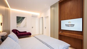 Quarto de hotel moderno com cama, uma cadeira vermelha, uma TV exibindo 'Grandstay Collection' e um guarda-roupa de madeira.