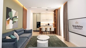 Uma sala moderna com sofá, mesa de centro, área de jantar com cadeiras, cozinha ao fundo e um letreiro Grandstay Collection na parede.