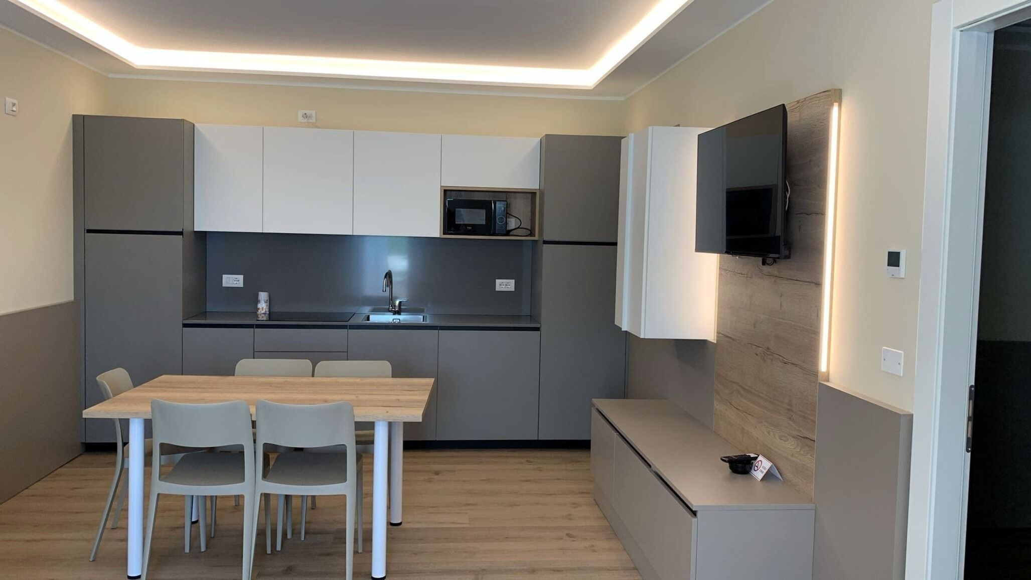 A modern kitchen with grey cabinets, a microwave, and a sink. There is a dining table with chairs and a bench. A flat-screen TV is mounted on the wall.