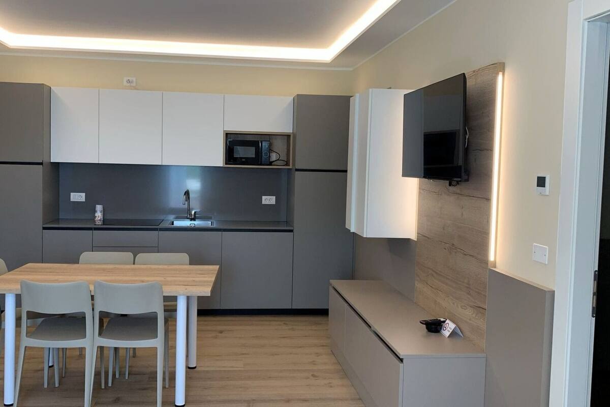 A modern kitchen with grey cabinets, a microwave, and a sink. There is a dining table with chairs and a bench. A flat-screen TV is mounted on the wall.