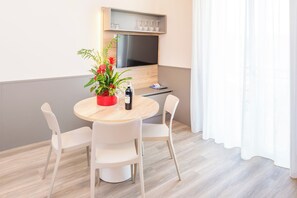 Une pièce moderne avec une table ronde en bois, des chaises blanches, une plante en pot, une bouteille et un téléviseur à écran plat fixé au mur.