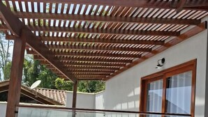 Un espacio exterior cubierto con una pérgola de madera, barandilla de vidrio y una escalera que lleva a la entrada de un edificio.