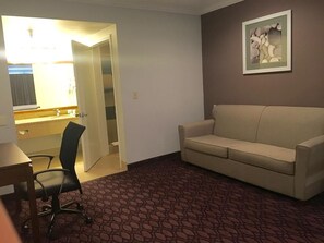 A hotel room with a beige sofa, a desk with a chair, a bathroom with a sink and mirror, and a framed picture on the wall. - Crimson Hotel (Manhattan Beach)