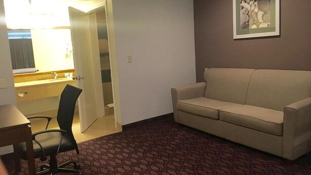 A hotel room with a beige sofa, a desk with a chair, a bathroom with a sink and mirror, and a framed picture on the wall. - Crimson Hotel (Manhattan Beach)