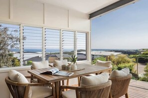 A wooden table with chairs, a book, and a potted plant on a balcony with a view of the ocean and buildings.