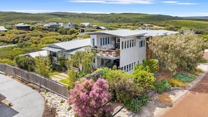 A modern two-story house with a balcony, surrounded by a landscaped garden and a view of rolling hills.