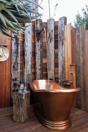 A rustic outdoor setting with a copper bathtub, wooden fence, and a small wooden stool.
