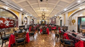 Una àmplia sala de dinar amb alts sostres i penjolls ornamentals, amb diverses taules preparades, cobertes amb teles vermelles i cadires de fusta.