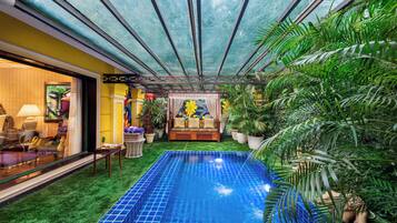A pool area with a glass ceiling, surrounded by lush greenery and a sofa with cushions.