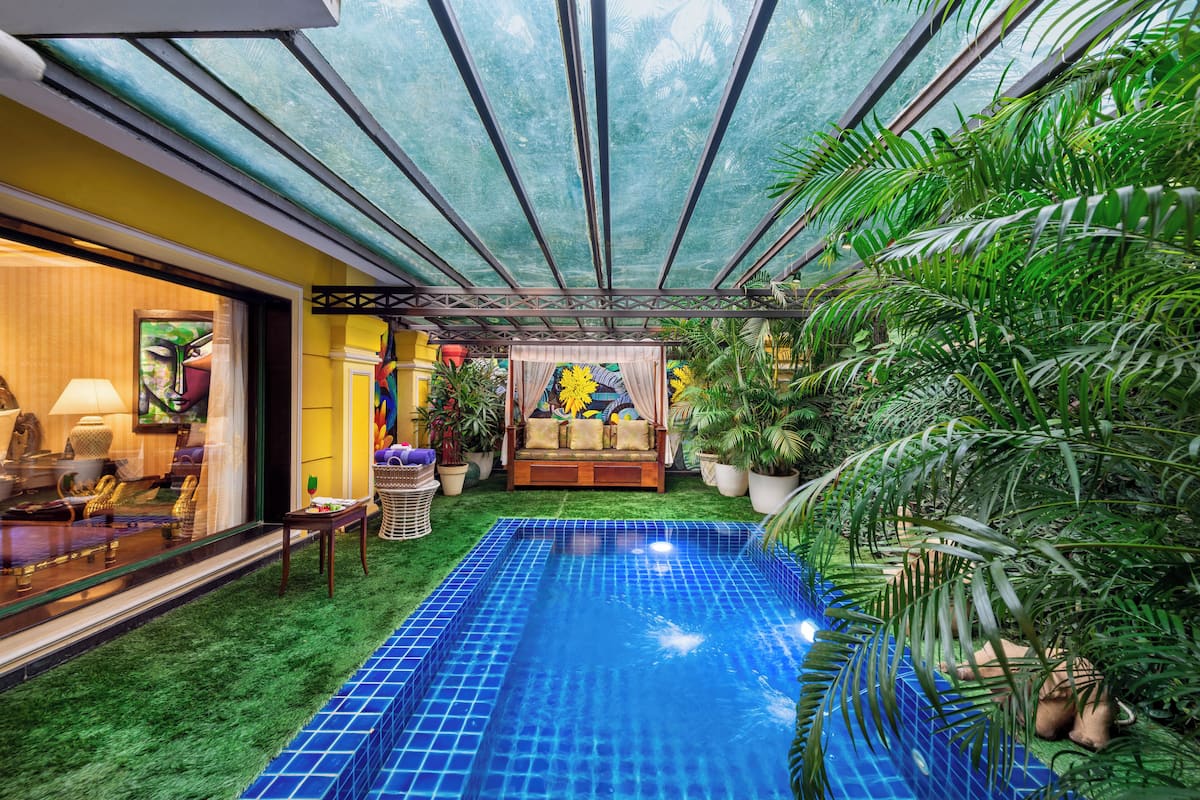 Un espace piscine doté d’un toit en verre, entouré d’une végétation luxuriante et agrémenté d’un canapé avec des coussins.