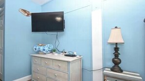 A room with a flat-screen TV mounted on the wall, a dresser with drawers, a lamp, and a small table with a lamp. - Cedar Key Turtle 132 (Cedar Key)