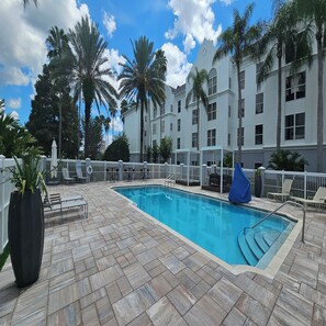 A swimming pool with a tiled deck, palm trees, and a white building with multiple windows. - Revival Scottsdale Studios (Orlando)