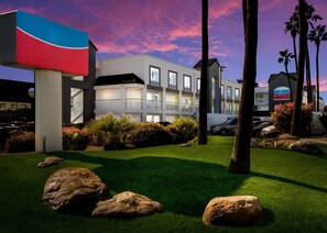 Un hotel de varios pisos con diseño moderno, con balcones y un jardín ajardinado con rocas y palmeras.