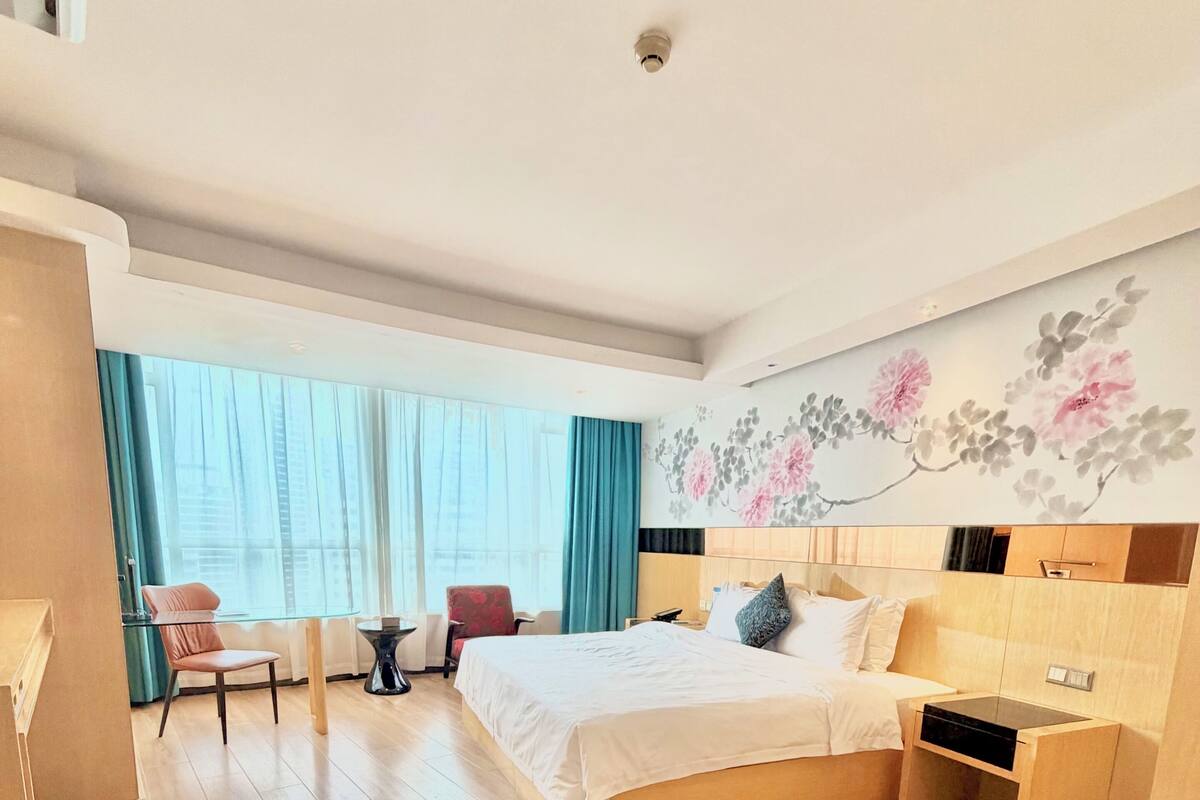 Ein Hotelzimmer mit Bett, Schreibtisch, Stuhl, einem Fenster mit Vorhängen und einer Wanddekoration mit floralem Muster.