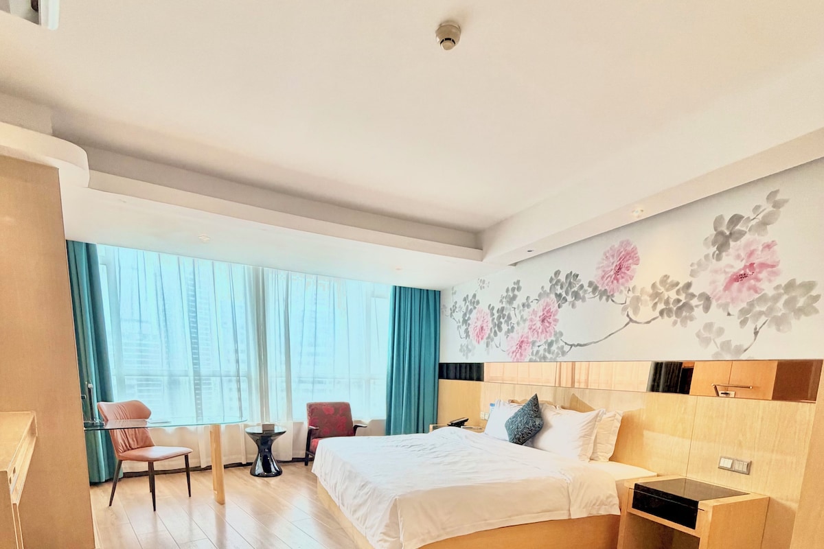 Ett hotellrum med en säng, ett skrivbord, en stol, ett fönster med gardiner och en blommig väggdekoration.