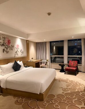 Ett hotellrum med en stor säng, ett skrivbord, en stol och utsikt över staden på natten.