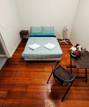 A small room with a bed, a chair, a small table, and a fan. - Surf 'N' Snow Backpackers (Auckland)
