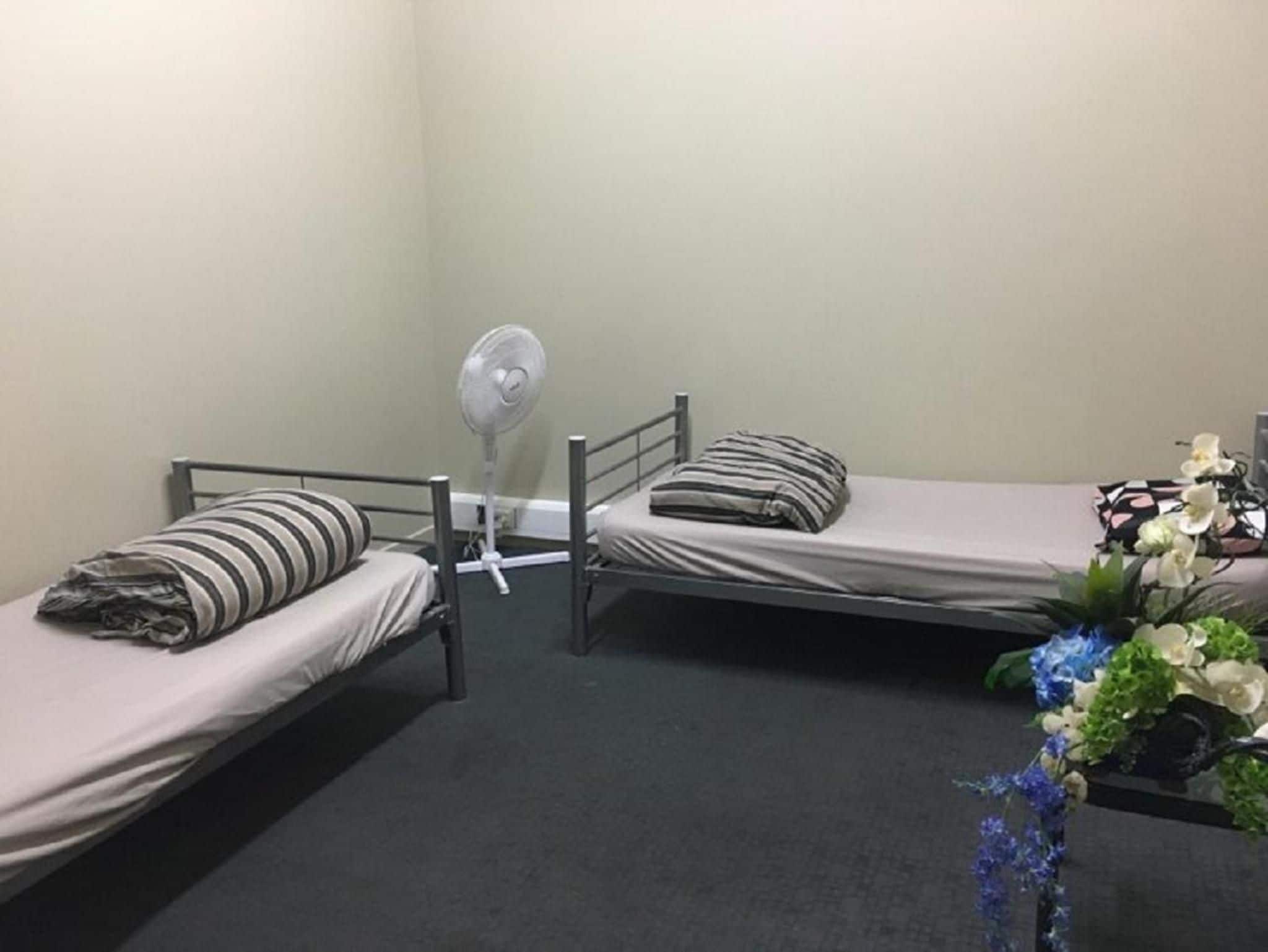 A small, single-room accommodation with two single beds, a standing fan, and a small table with a vase of flowers.