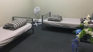Eine kleine Einzelunterkunft mit zwei Einzelbetten, einem Standventilator und einem kleinen Tisch mit Blumen.'