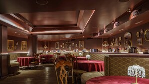Un restaurante tenuemente iluminado con manteles rojos, sillas de madera y una zona de bar con libros y cuadros enmarcados.