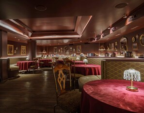 Ein gedämpft beleuchtetes Restaurant mit roten Tischdecken, Holzsesseln und einer Bar mit Büchern und gerahmten Bildern.