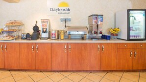 Ein Frühstücksbuffet mit einem Schild, auf dem 'Daybreak Breakfast' steht, einem Waffeleisen, einer Kaffeemaschine, einem Toaster und einem Kühlschrank mit dem Aufkleber 'Cold Drinks'.