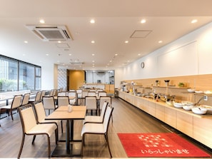 A modern dining area with tables and chairs, a kitchen in the background, and a red rug with Japanese characters. - APA Hotel Marugame-ekimae-odori (Marugame)