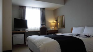 Ett hotellrum med en säng, ett skrivbord med en tv, ett litet kylskåp, en skrivbordslampa och ett fönster med gardiner.