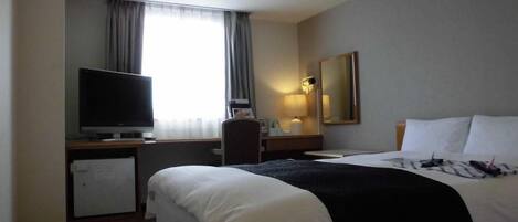 A hotel room with a bed, a desk with a television, a small refrigerator, a desk lamp, and a window with curtains.